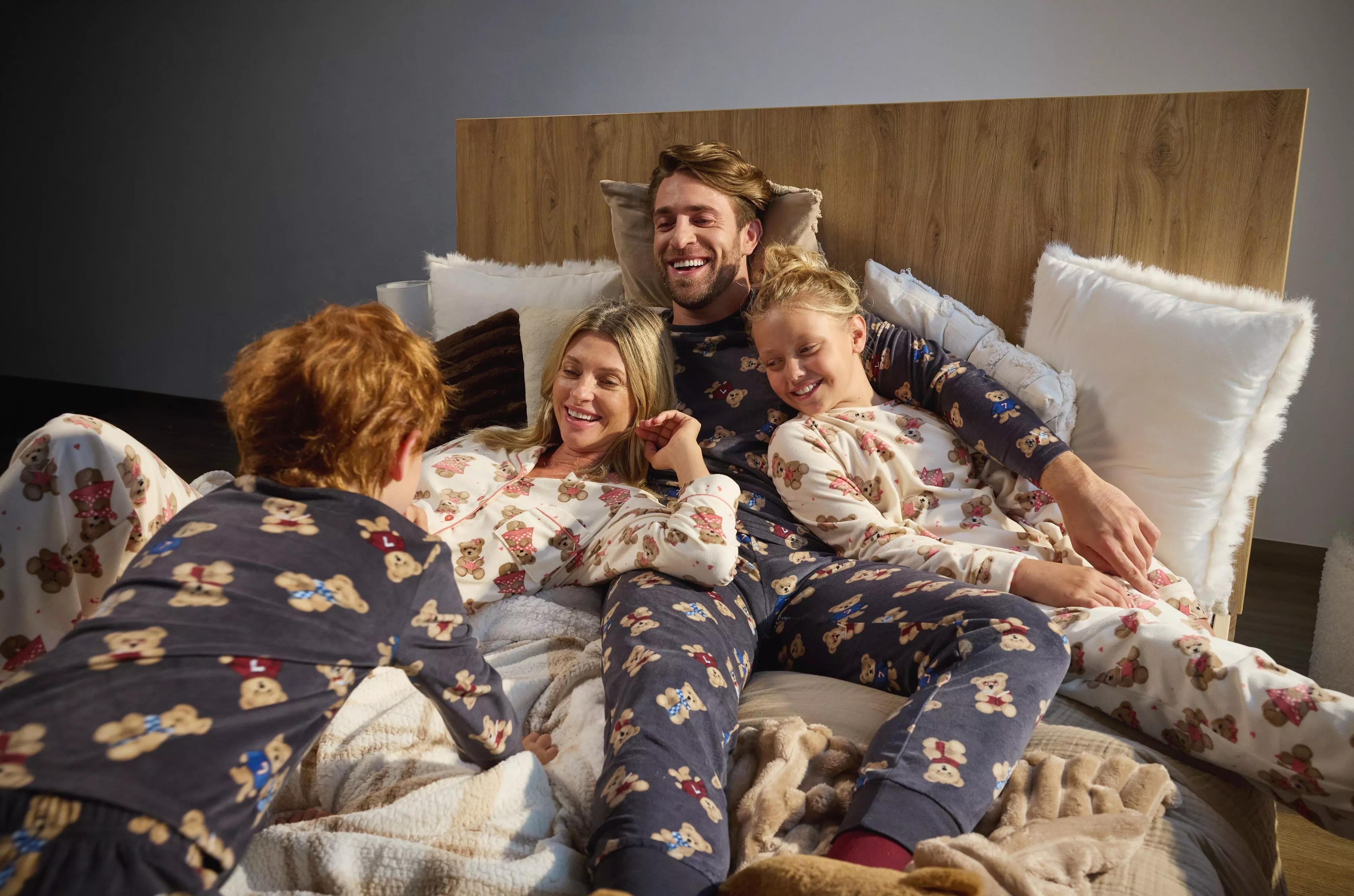 Pepco family of four relaxing together on bed wearing matching festive pyjamas with teddy bear and gingerbread prints.